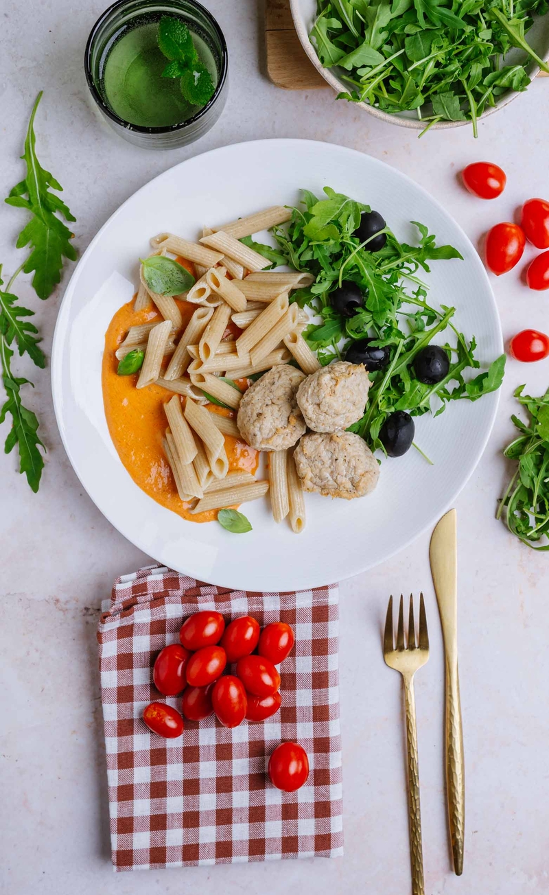 Pełnoziarnisty makaron penne w kremowym sosie pomidorowym z drobiowymi pulpecikami, rukola i czarne oliwki
